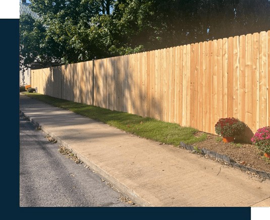 A wooden fence, installed by a skilled fencing installer in Central PA, lines a sidewalk bordered by grass and potted flowers, with trees visible in the background.