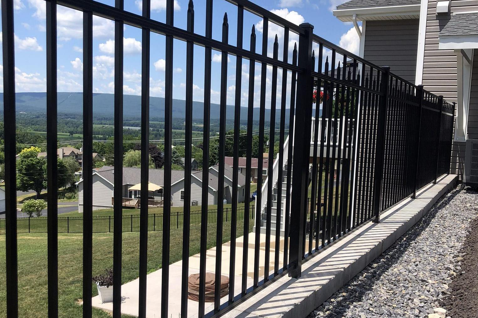 A black metal fence on a concrete base offers a stunning view of the residential neighborhood, with distant hills under a blue sky. Expertly crafted by a top fencing installer in Central PA, it seamlessly blends durability with scenic beauty.